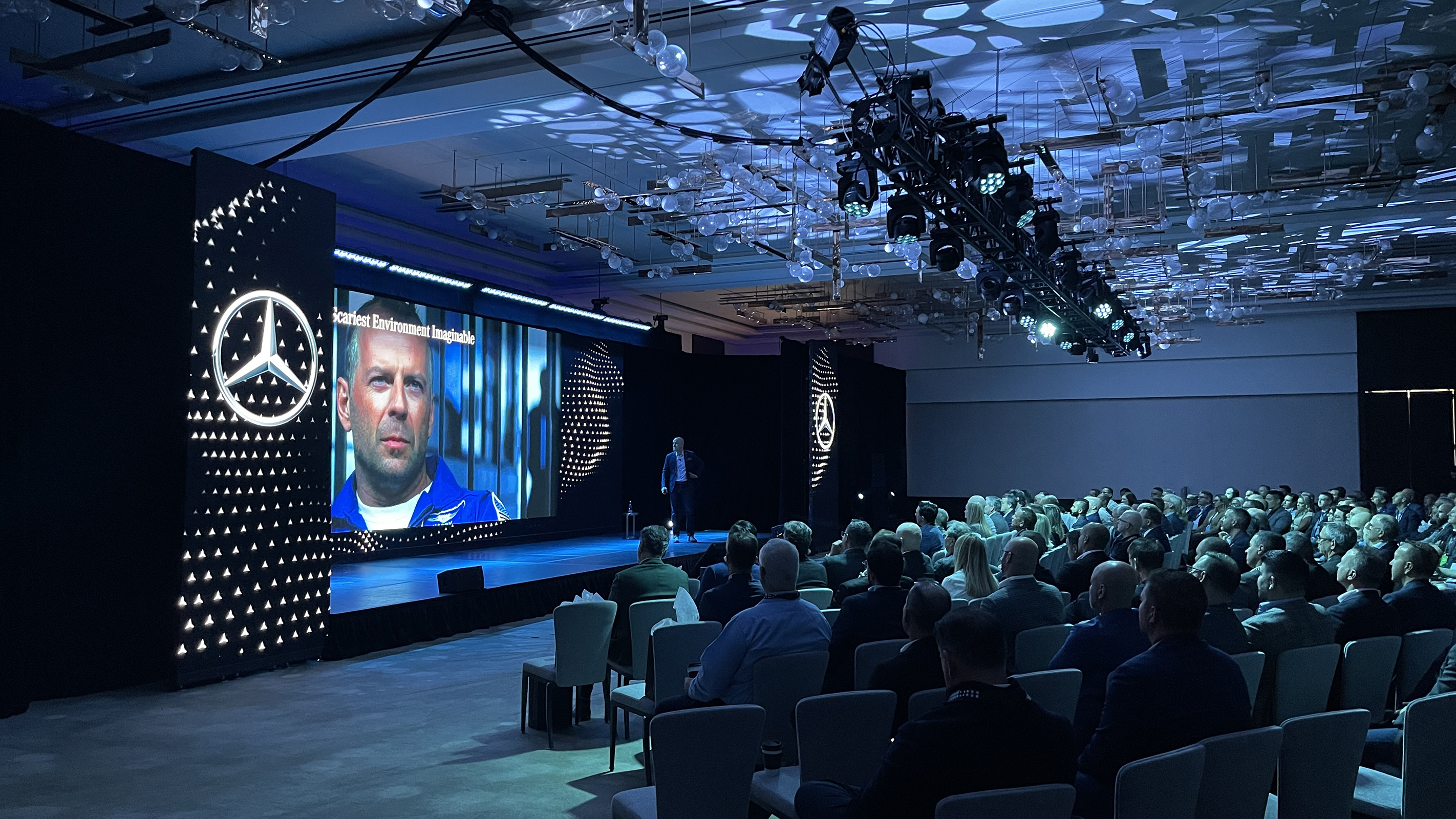 A large audience watches a presentation in a modern, dimly lit conference room. The stage features the Mercedes-Benz logo, colorful lights, and a large screen displaying a man's face and text.