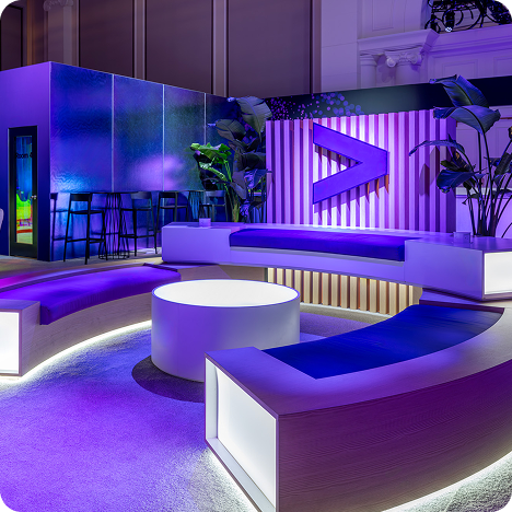 A modern lounge with curved seating, illuminated in purple and blue light, features a round table and a large chevron wall design—perfect for CES AV production events. Tall plants and a stylish bar area are visible in the background.