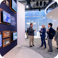 Three people stand and talk in front of a wall displaying multiple framed screens at a modern, well-lit CES AV production exhibition or trade show with blue and white decor.
