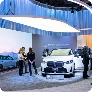 A group of people stand near a white BMW car on display at an indoor auto show, surrounded by CES AV production setups, with modern lighting, informational displays, and a large screen in the background.