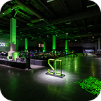 A large indoor event space illuminated with green lights, featuring computer stations and a glowing "SI" sign in the foreground. The modern, tech-focused atmosphere suggests high-end CES AV production.