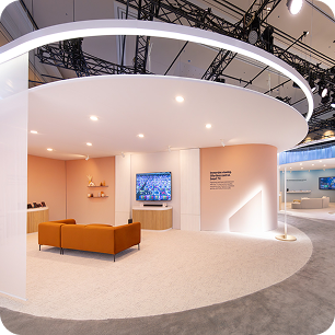 Modern exhibition booth with a circular white ceiling, soft lighting, orange sofa, wall-mounted TV, and minimalist decor on light carpet; open design ideal for CES AV production, with additional booths visible in the background.