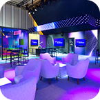 Modern lounge area with white armchairs, high tables, and multiple screens displaying “Welcome.” The space features CES AV production touches, brightly lit by purple and blue ambient lighting and colorful decor.