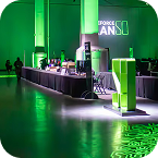 A modern indoor event space, designed with CES AV production quality, is lit with green lights and features tall pillars, a long counter displaying items, and a projection on the wall reading “STORAGE CAN.”.