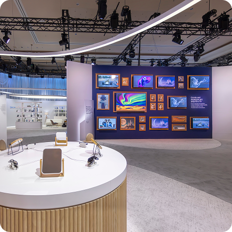 A modern exhibition space with CES AV production, featuring digital screens displaying colorful artwork on a curved wall, interactive tables with tablets and headphones in the foreground, and sleek lighting overhead.