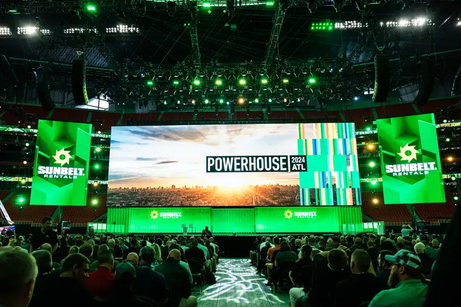 A large crowd faces a brightly lit stage with screens displaying "POWERHOUSE 2024 ATL" and "Sunbelt Rentals" logos, set inside a stadium with green lighting effects.