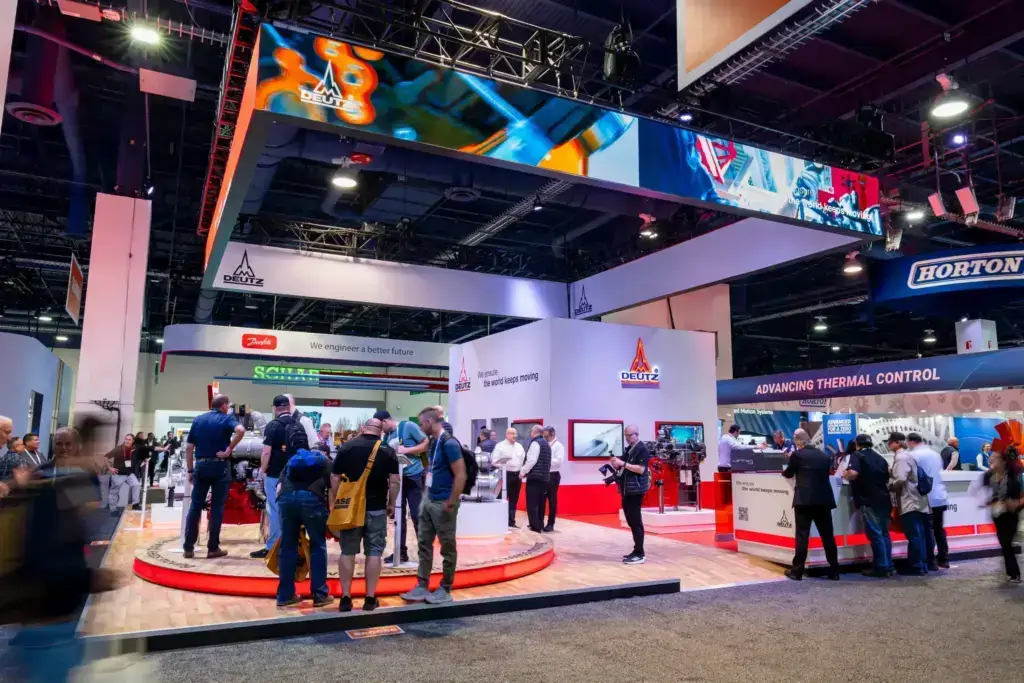 People gather at a brightly lit trade show booth with digital screens and displays. Several attendees take photos and talk, while branded signage and products are visible around the exhibit.