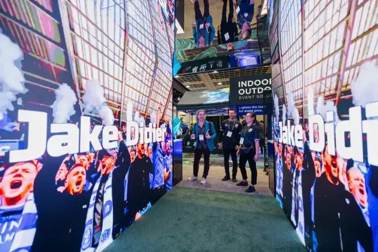 Three people stand in an event space, surrounded by large screens showing cheering fans and the partially visible text “Jake Didio.” The atmosphere is lively and modern.