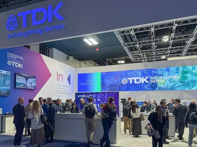 People gather at a TDK exhibition booth with blue and purple signage that reads “TDK In Everything, Better.” The booth features monitors and displays, with attendees interacting and networking.