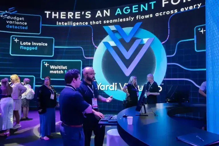 People interact at a tech expo booth with a large digital display reading “There’s an agent for that” and “Yardi Voyager.” Several participants talk and look at screens in a modern, blue-lit setting.