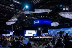 A large conference with many attendees seated around a central stage, large screens displaying “World,” and blue lighting overhead in a spacious, modern venue.