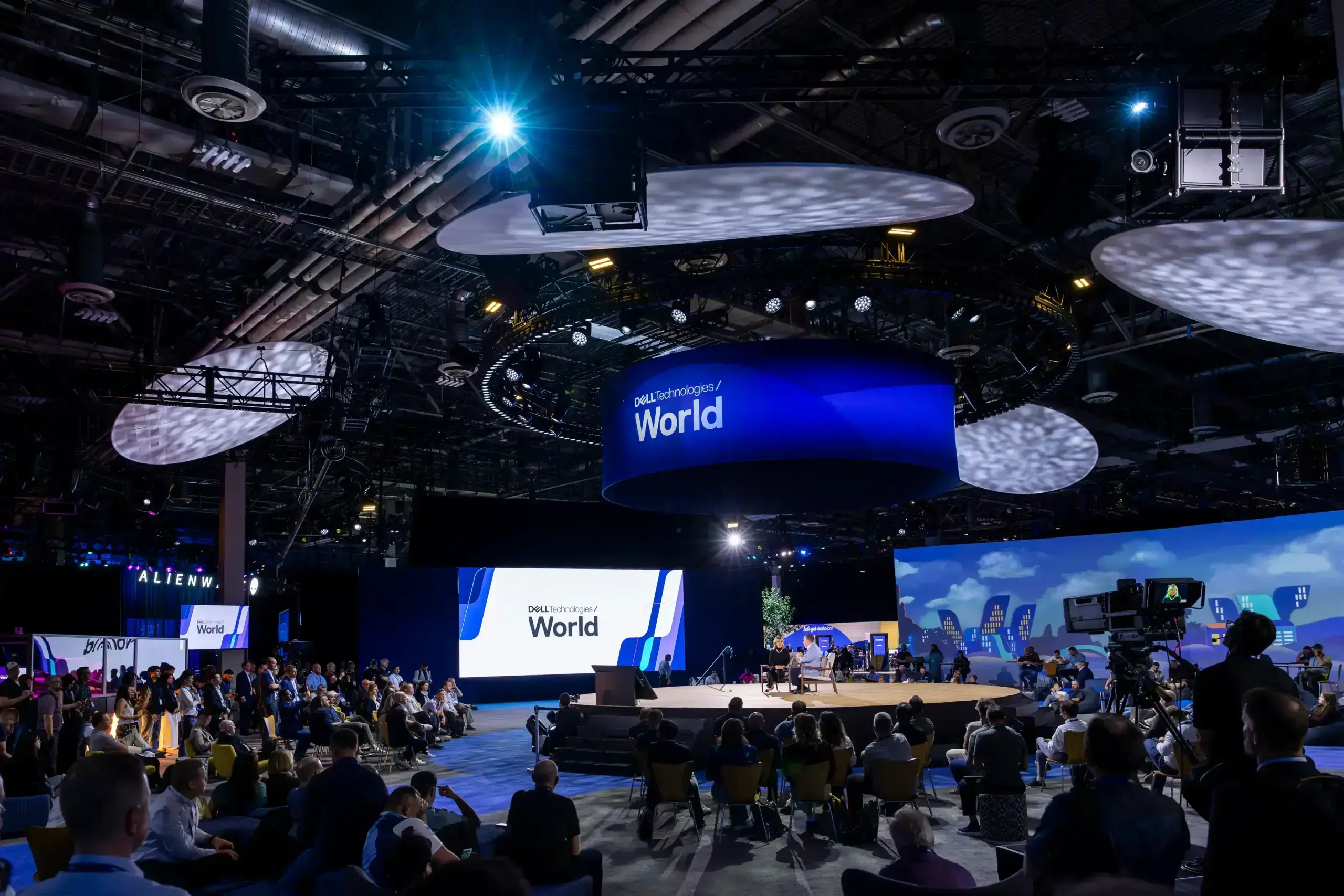 A large conference with many attendees seated around a central stage, large screens displaying “World,” and blue lighting overhead in a spacious, modern venue.