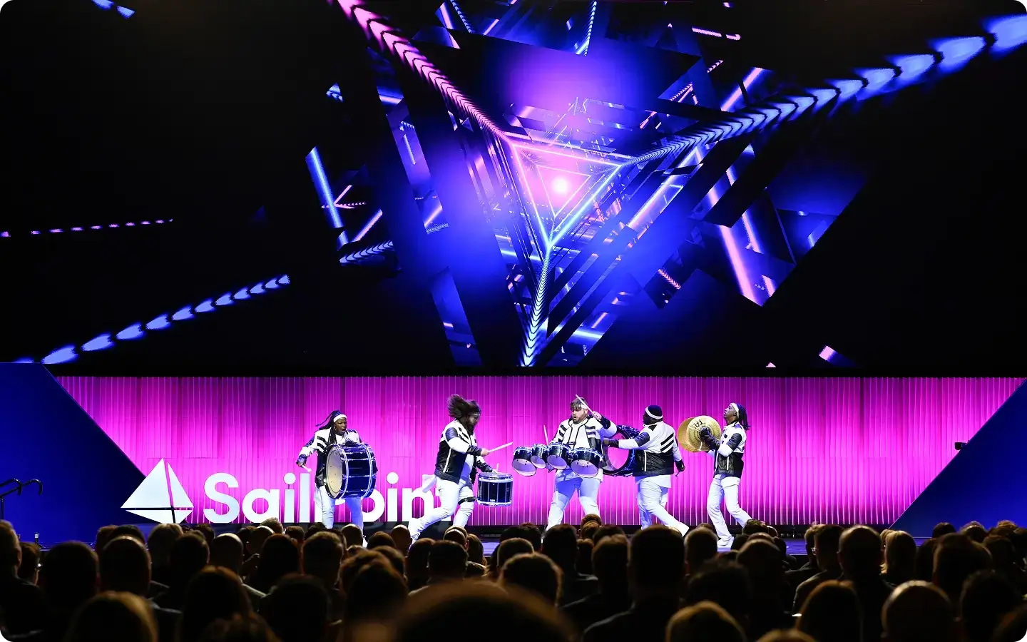 A drumming group performs onstage with purple and blue geometric lights and a SailPoint logo in the background, as an audience watches.